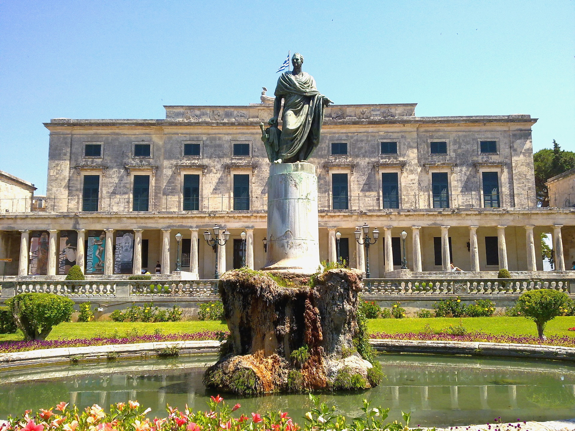 Fountain with statue of a man in a robe in the middle. Behind is a large building showing 18 columns in a row in the front of the building