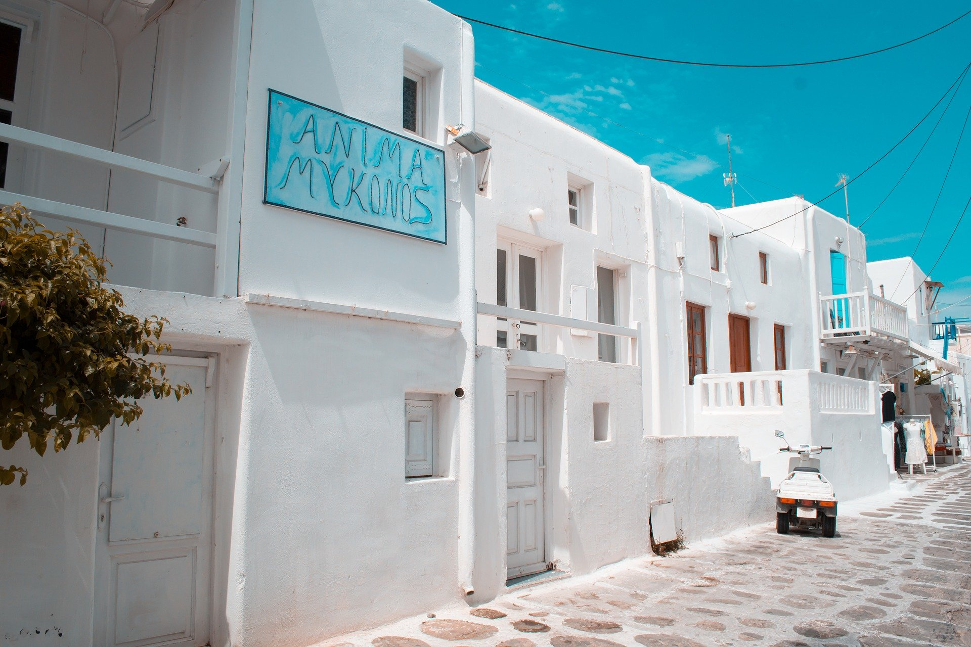 White buildings along a street, tan stone pavement. Blue sky, blue sign reading "Anima Mykonos"